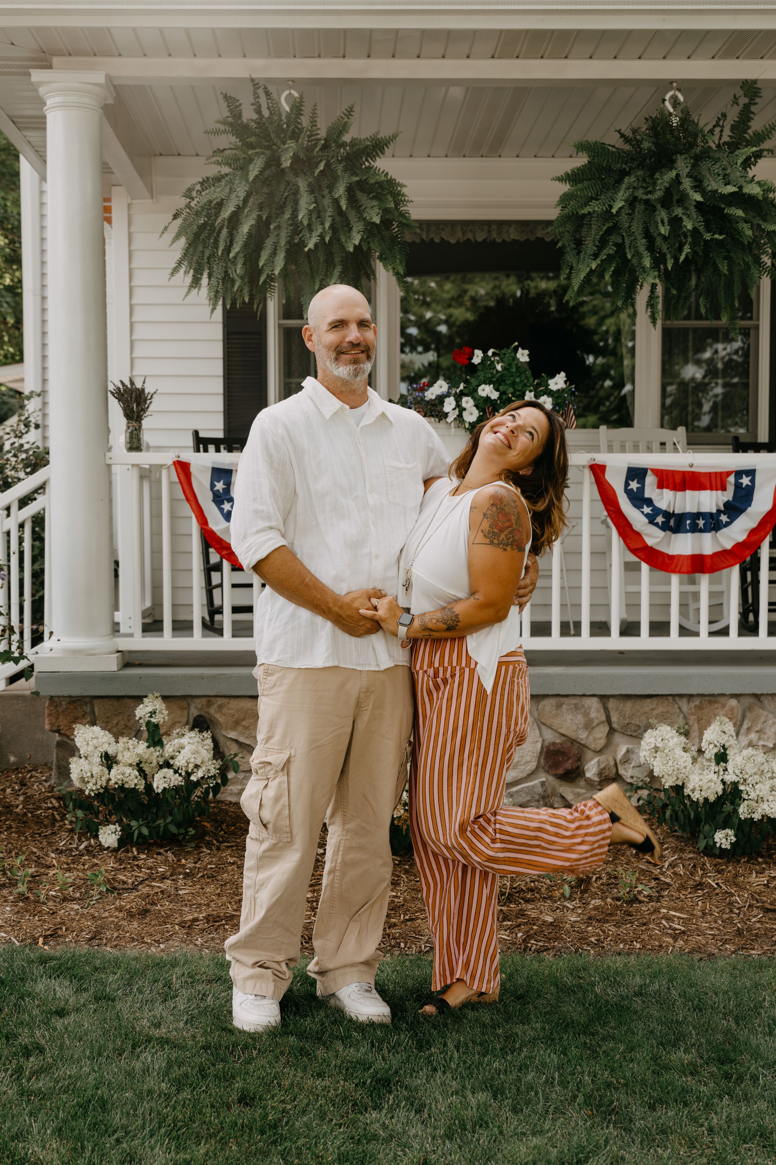 A romantic couple stands on a porch decorated with hanging plants and patriotic bunting, surrounded by blooming flowers.