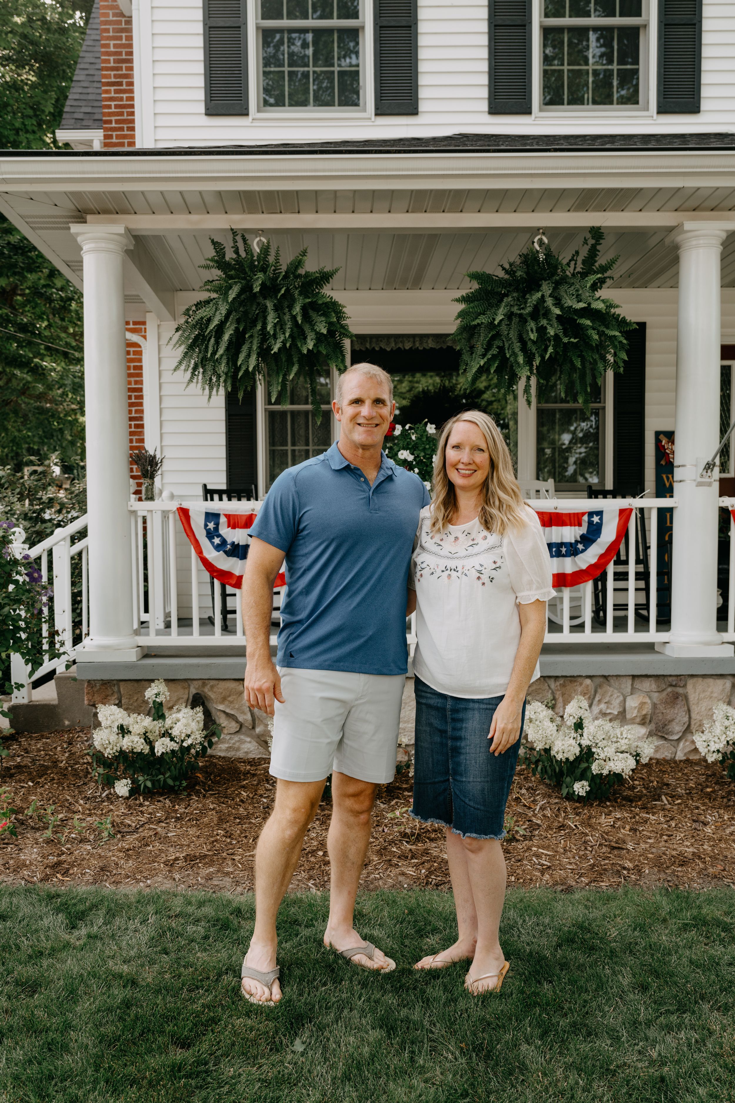 Family gathers on the front lawn of a classic white colonial home with patriotic bunting decorations.