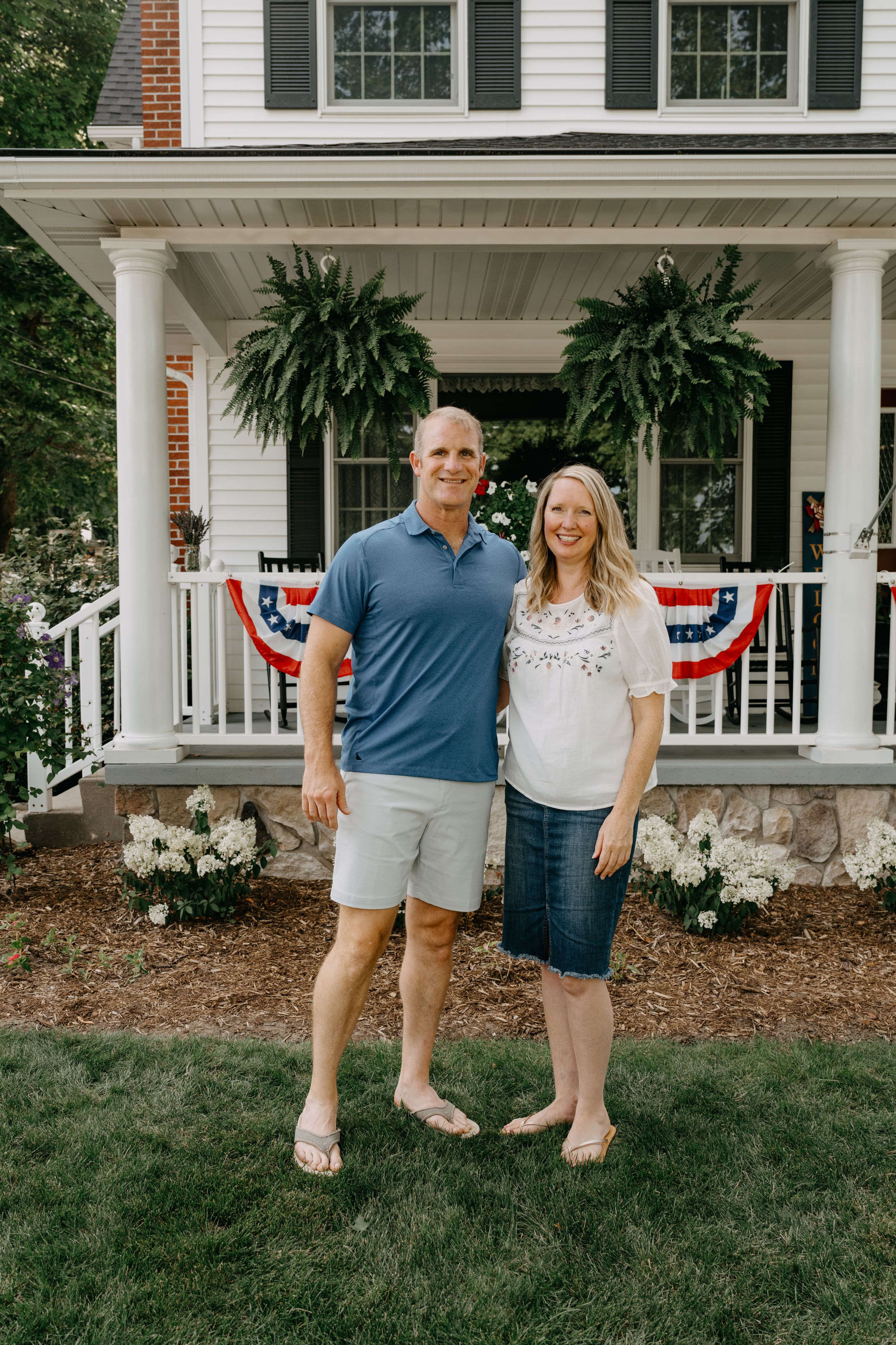 A couple stands on their front lawn in front of a white colonial porch decorated with ferns and patriotic bunting.