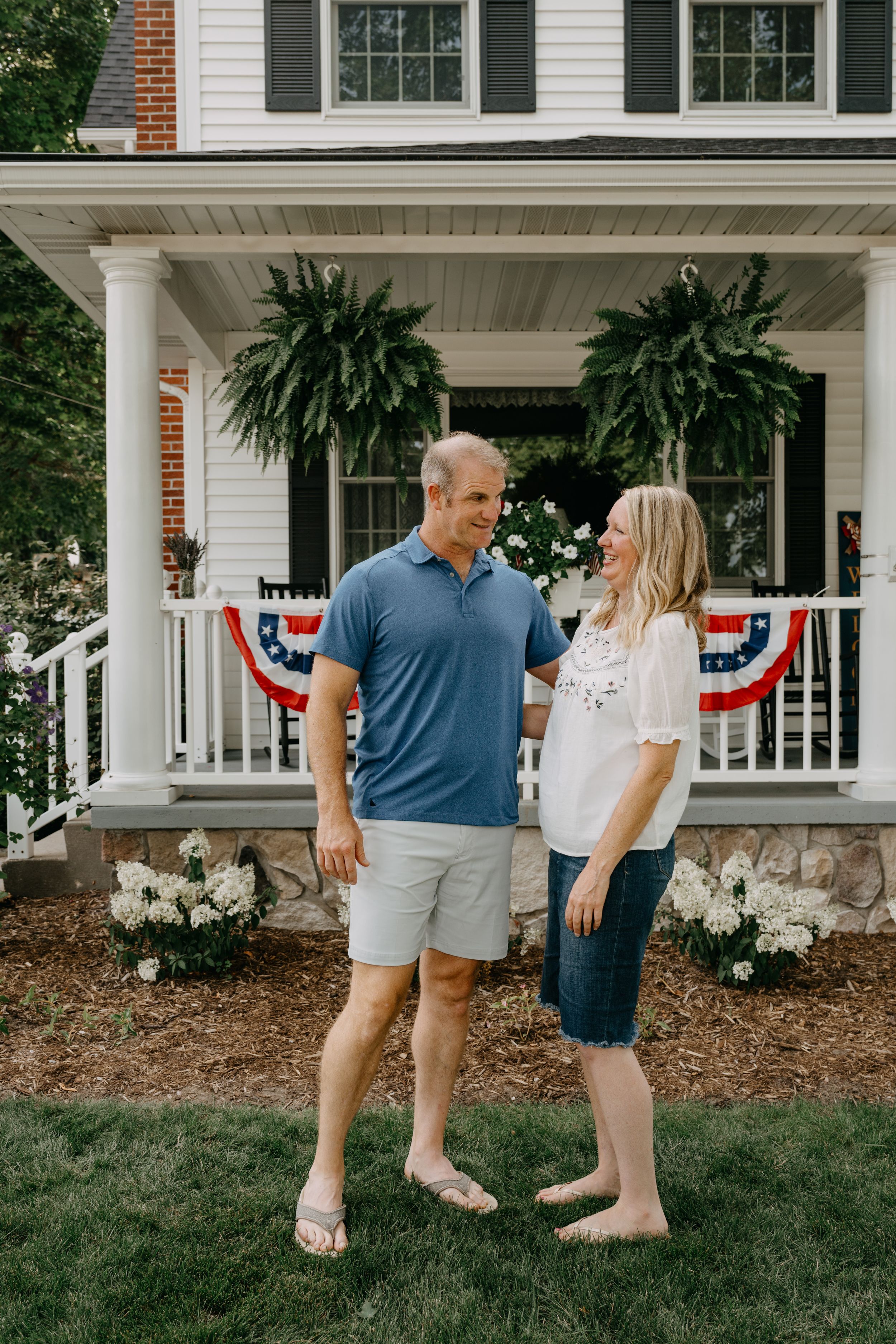 Casual couple stands barefoot on lawn in front of classic white porch decorated with patriotic bunting and ferns.