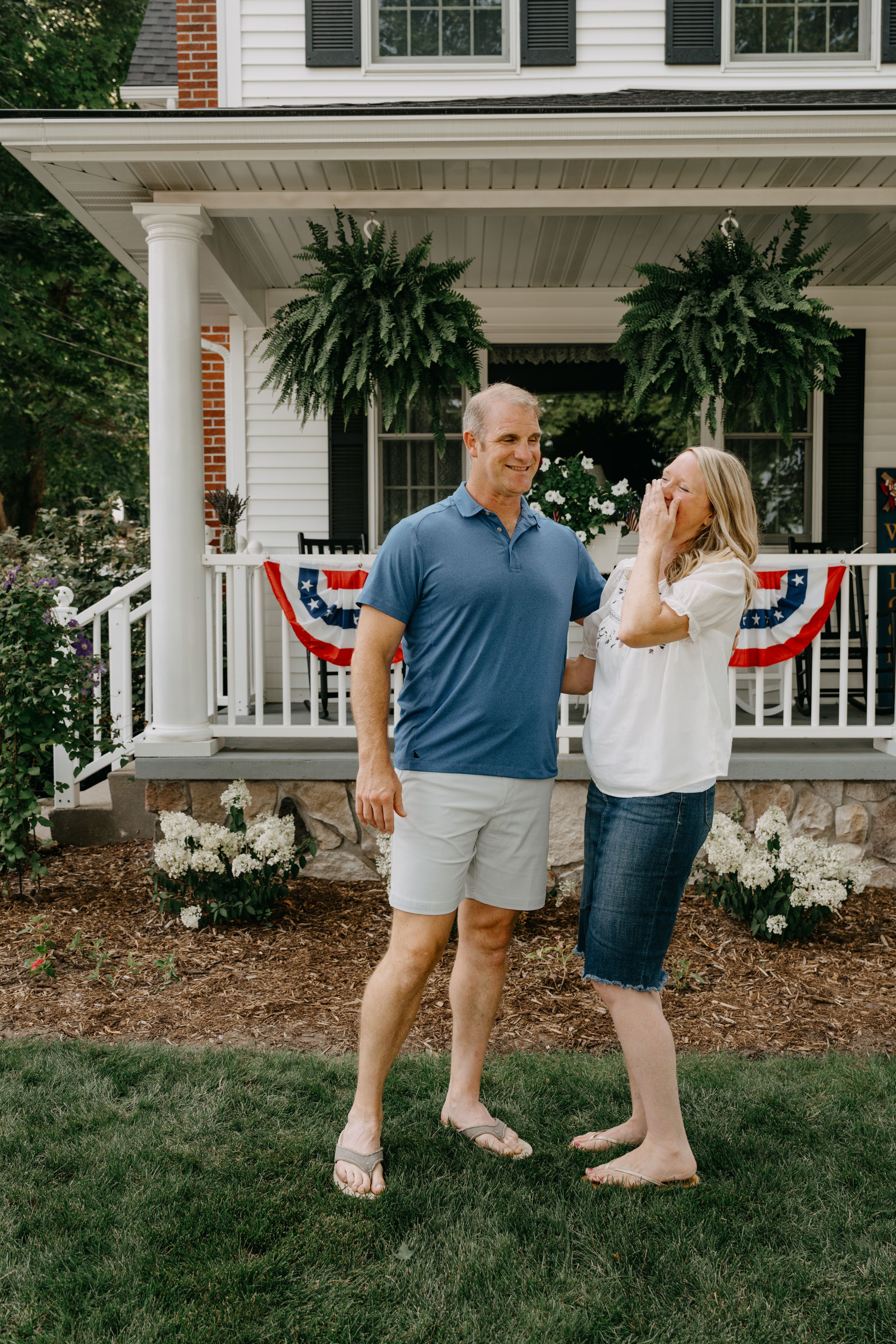 Couple stands on front lawn of white house with patriotic bunting and hanging ferns on porch during summer.