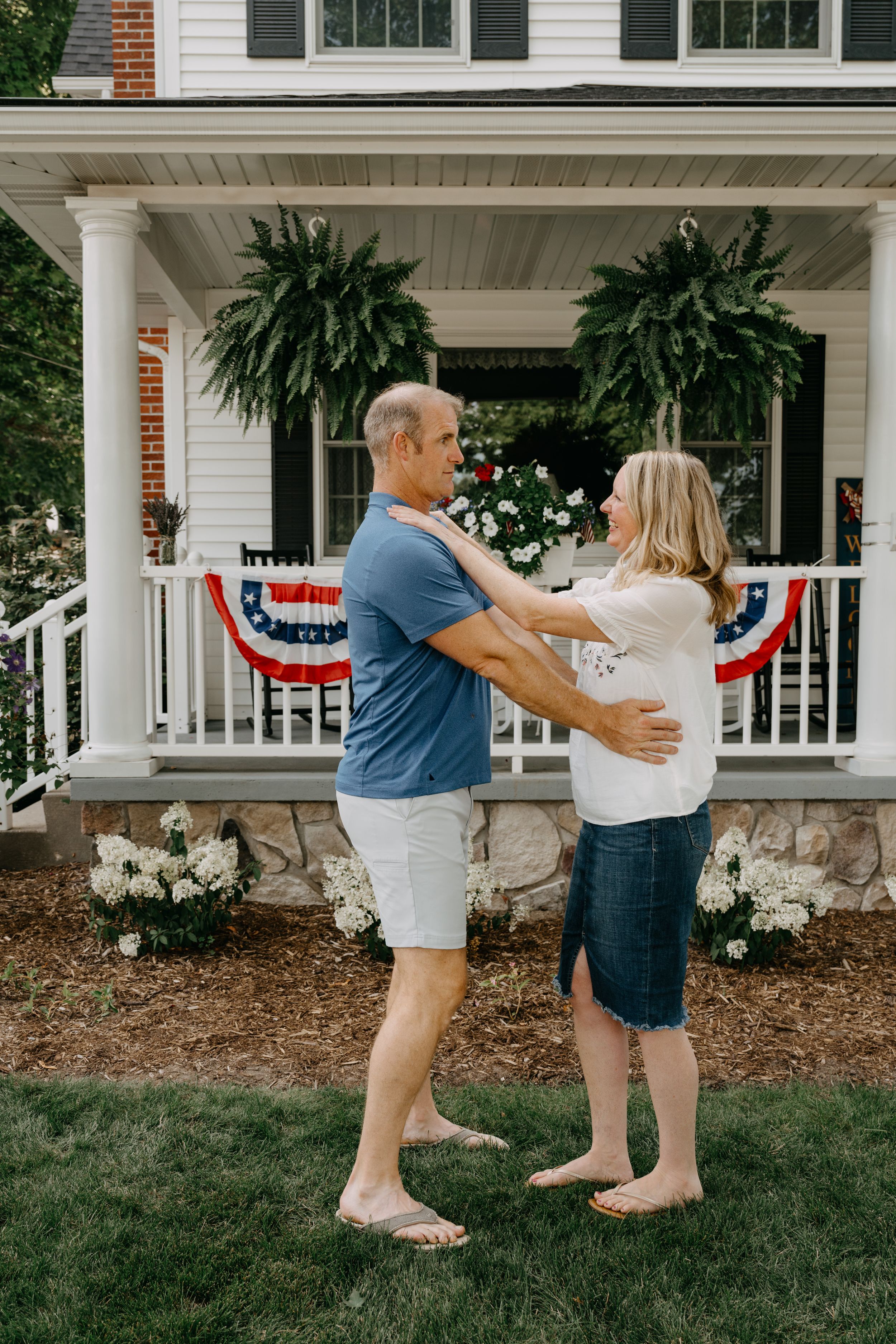 A couple dances together on the lawn in front of their porch adorned with American flag bunting and hanging plants.