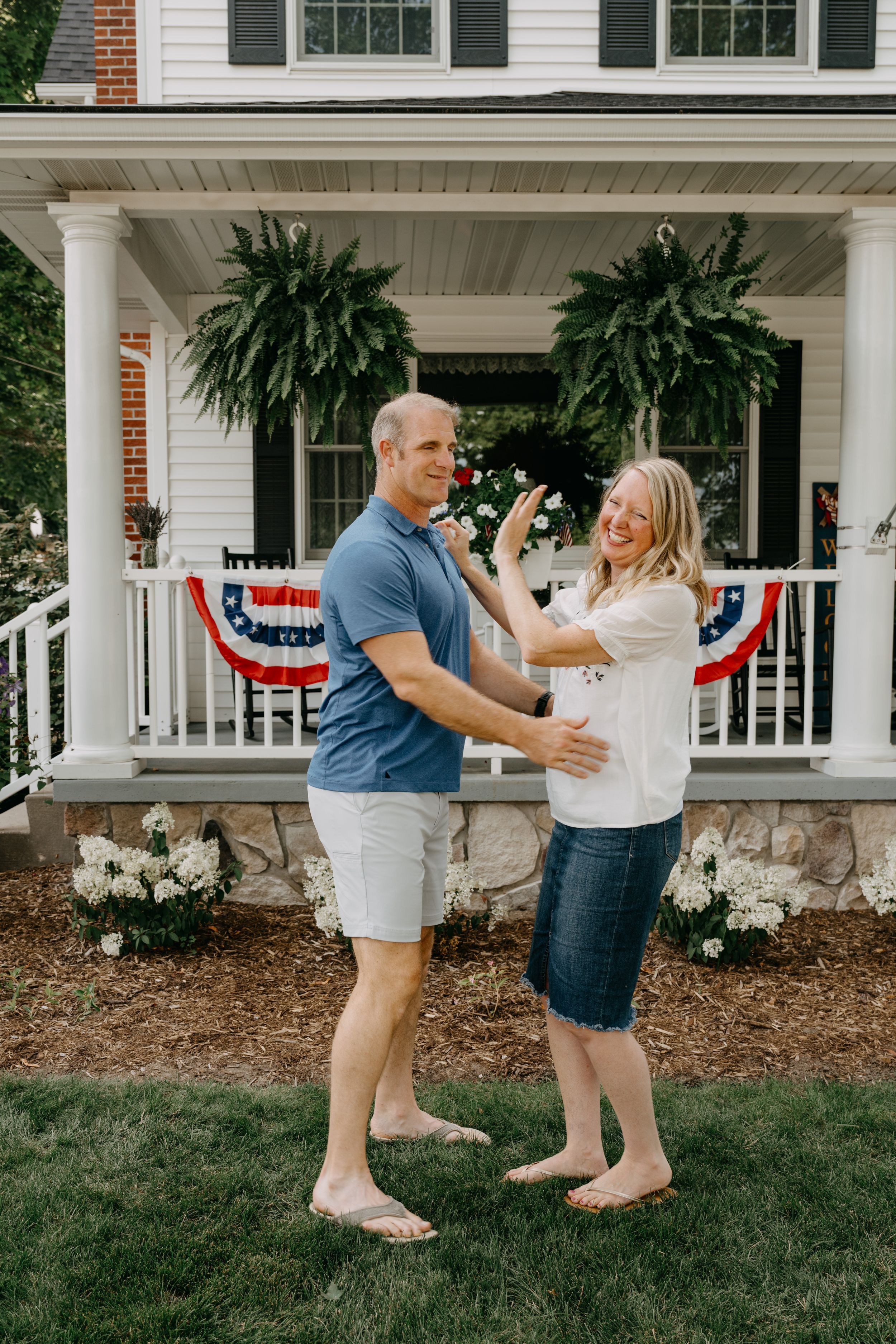 Happy couple shares a playful moment on their lawn in front of a patriotic-decorated white porch with ferns.