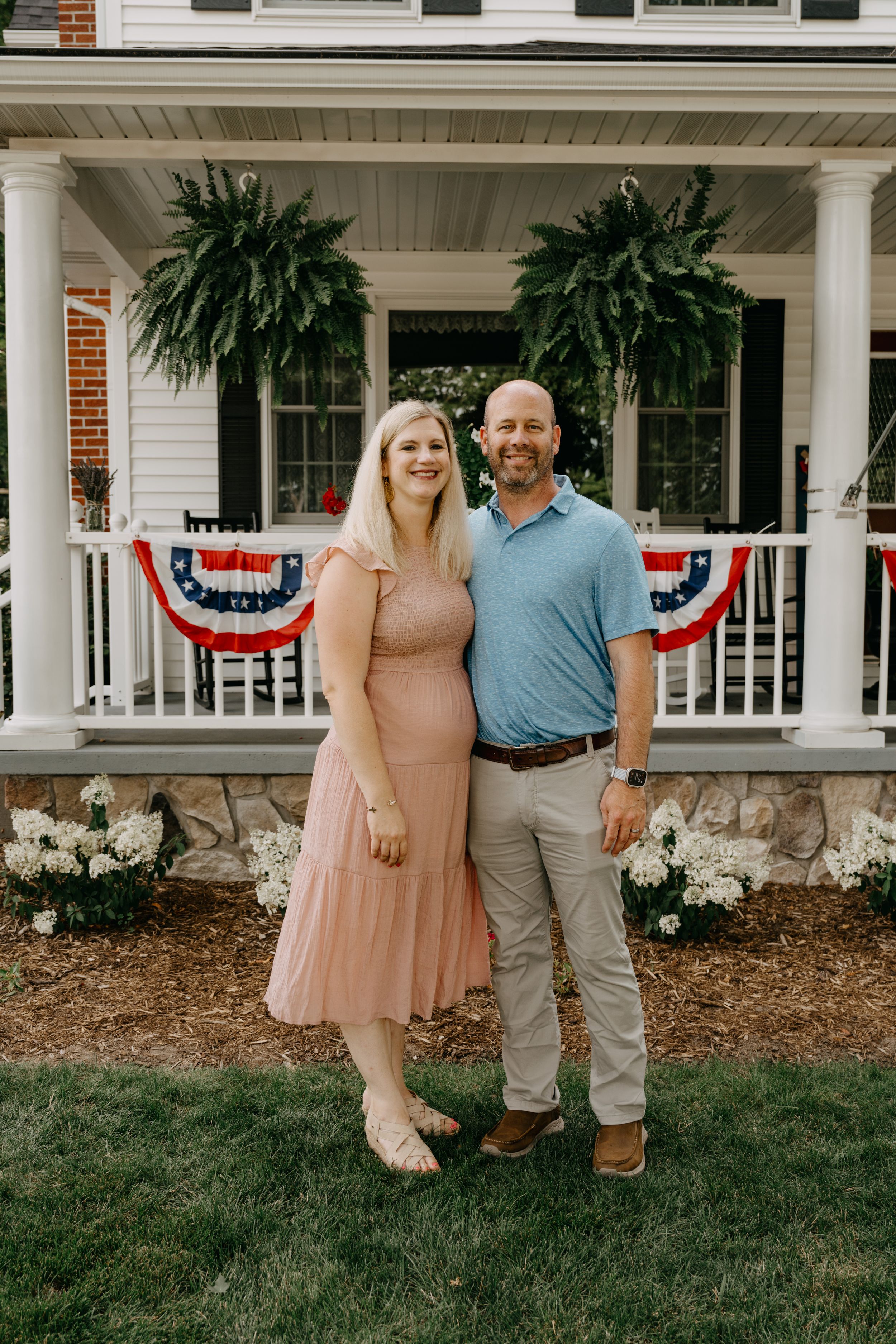 A couple stands together on their lawn in front of a charming white porch decorated with patriotic bunting and hanging plants.