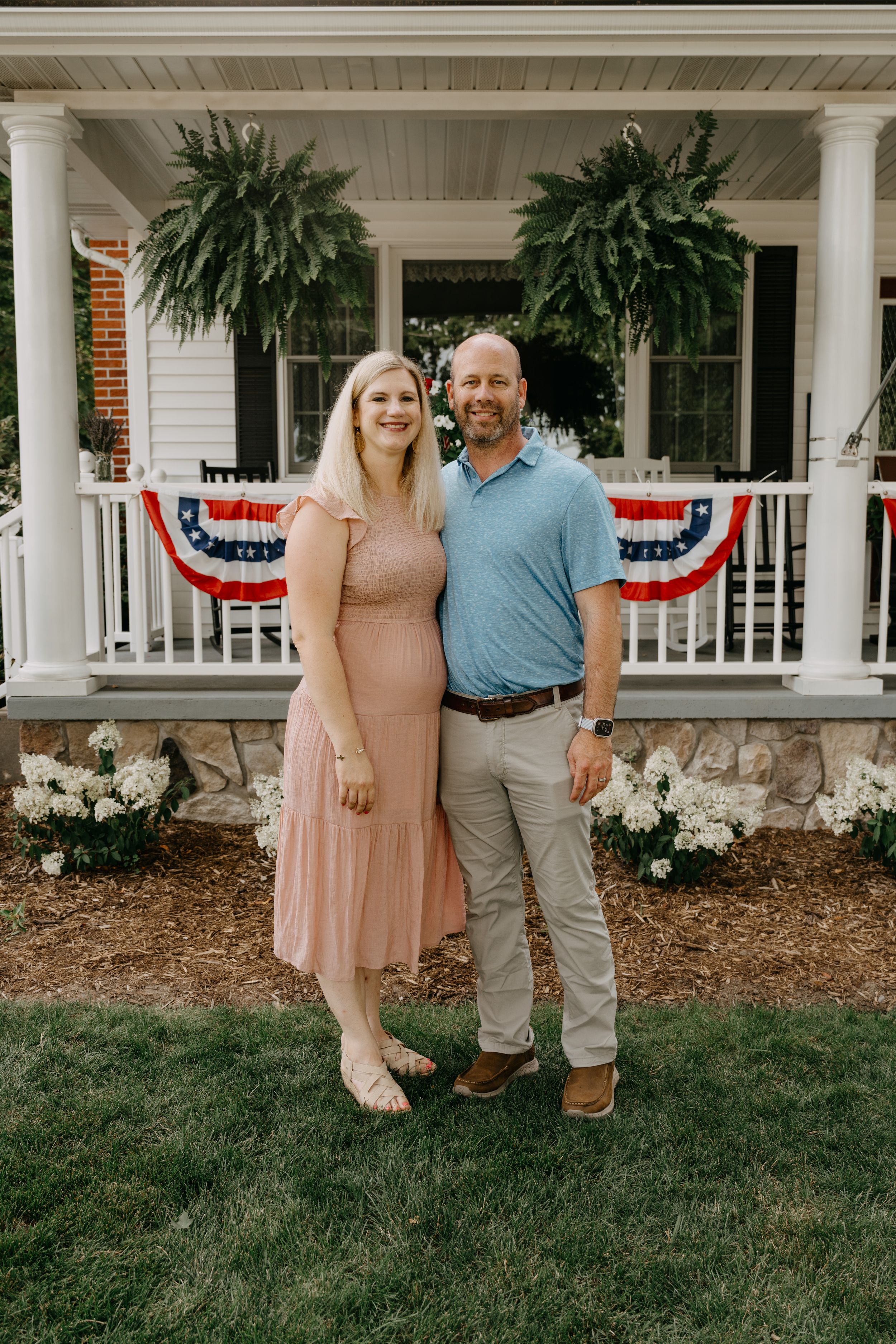 A couple poses on their front porch decorated with large ferns and patriotic bunting for summer.