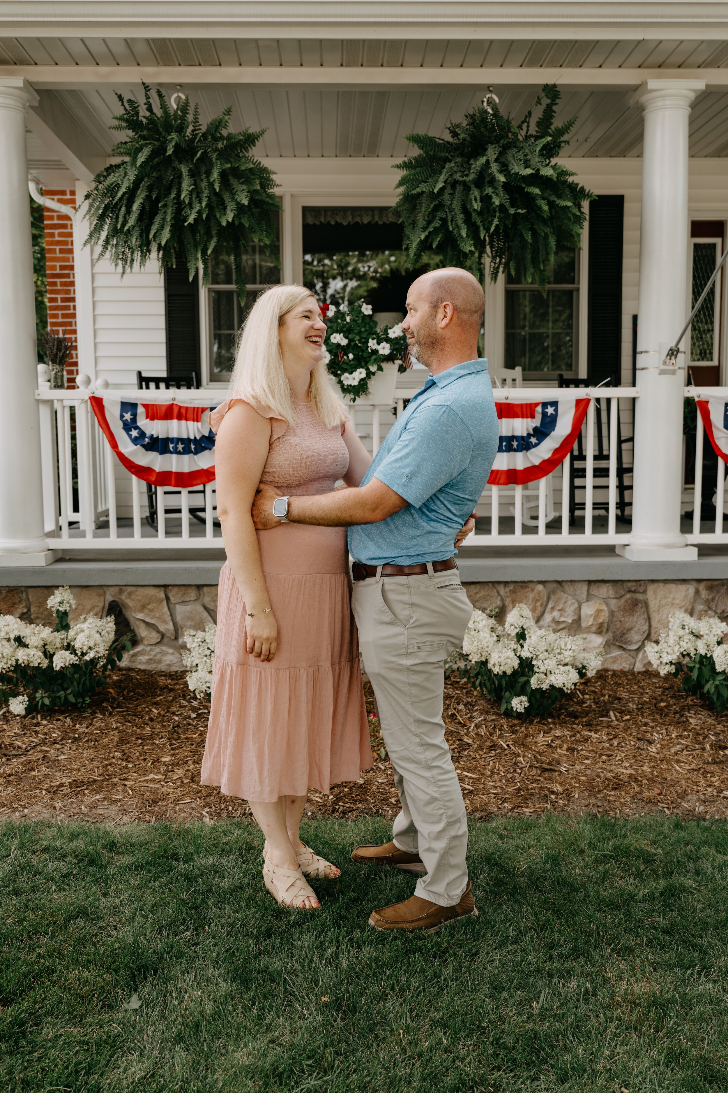 A couple embraces on a lawn in front of a porch decorated with patriotic bunting and hanging ferns.