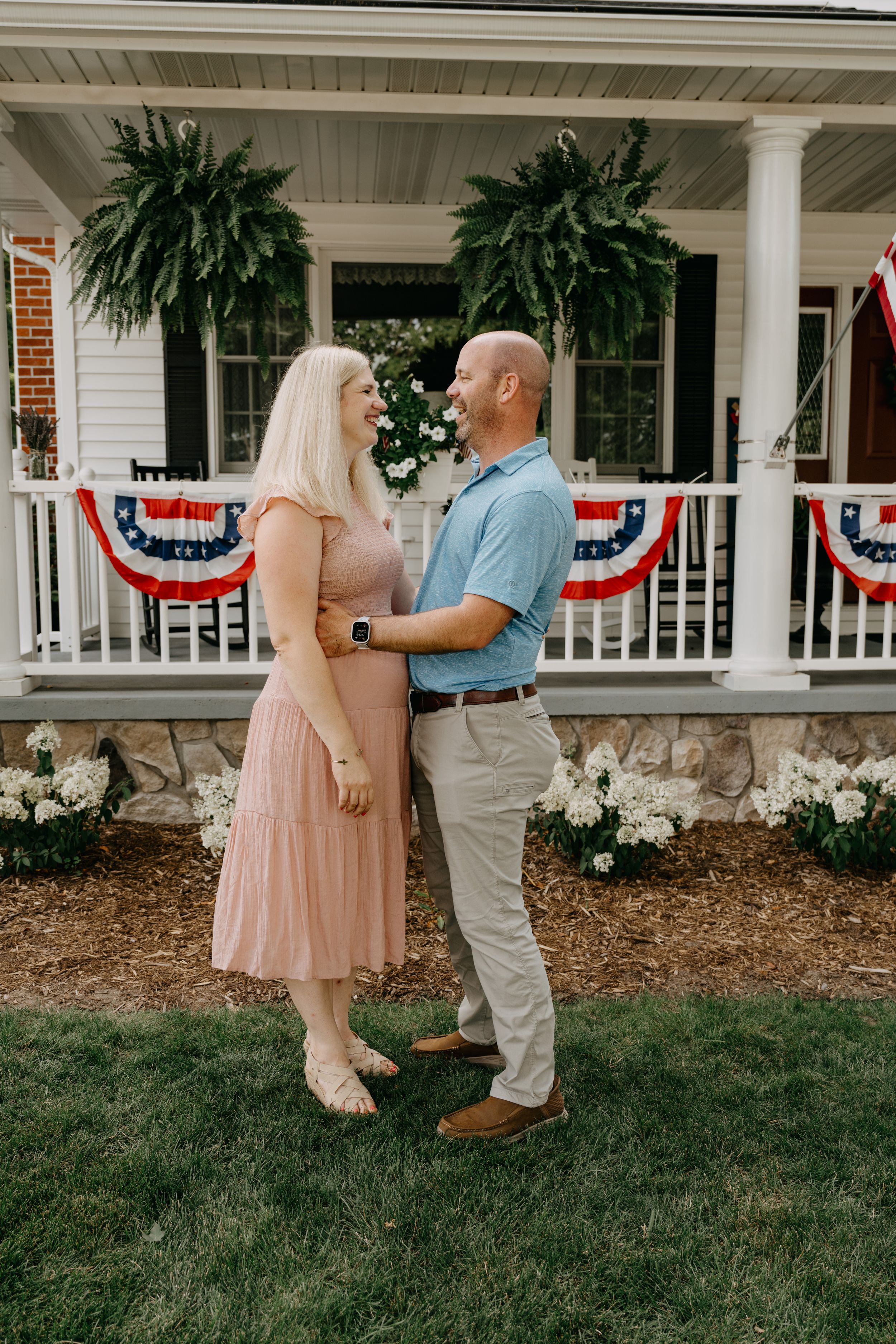 A couple stands on a front porch decorated with patriotic bunting and large hanging ferns.