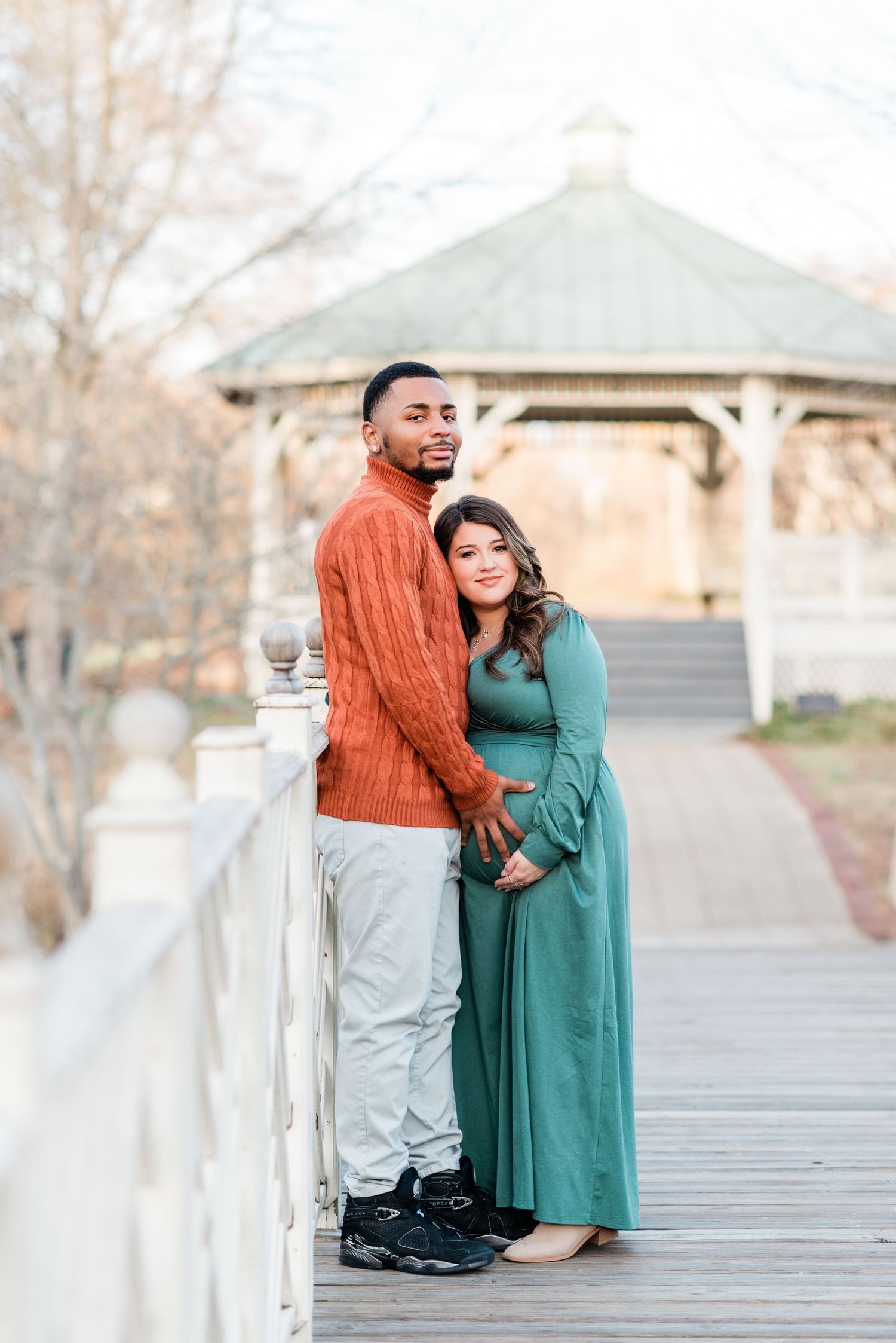 A couple in autumn attire stands on a wooden bridge near a gazebo on a crisp fall day.