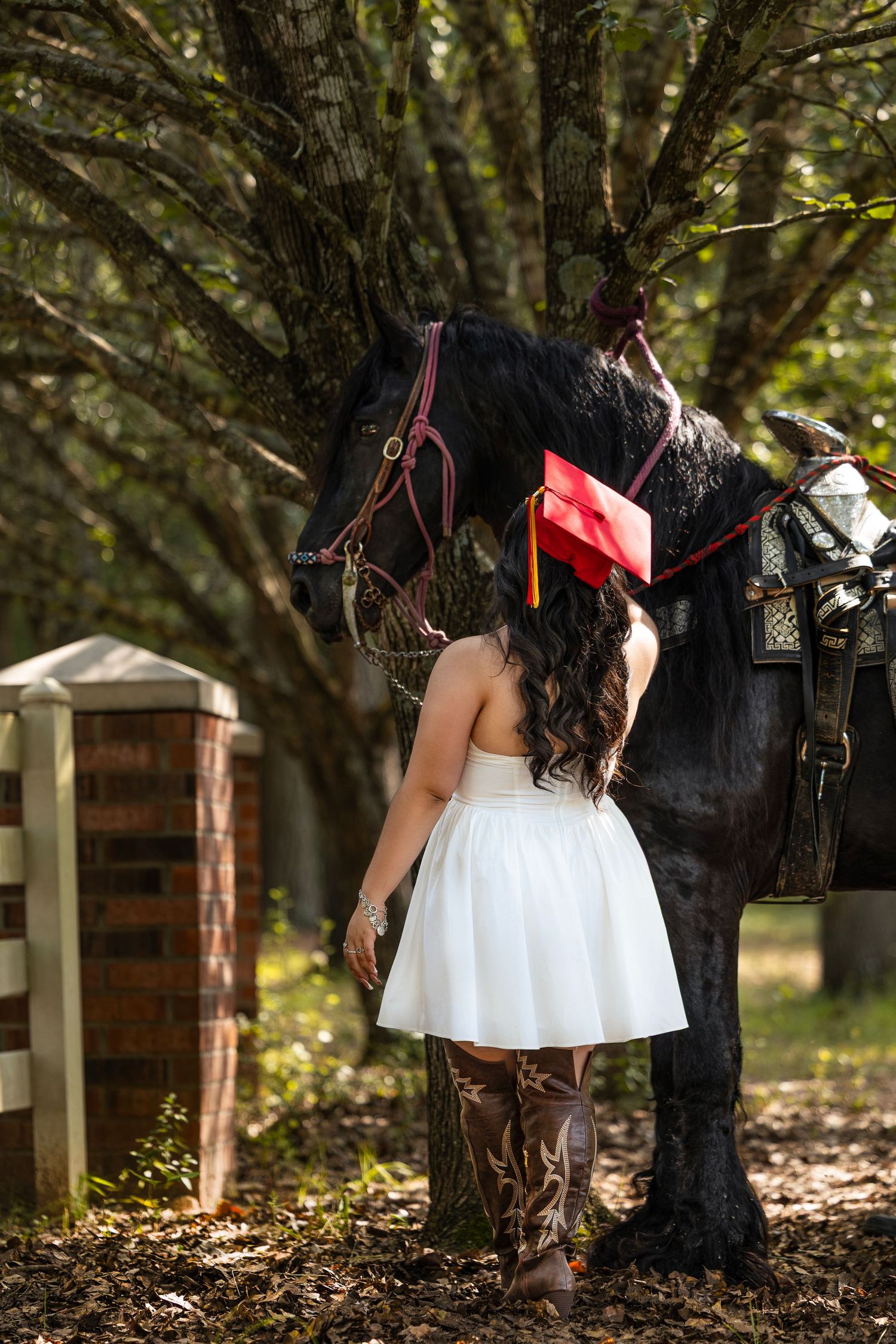 Graduate in white dress and red cap and gown poses outdoors against trees in natural lighting.