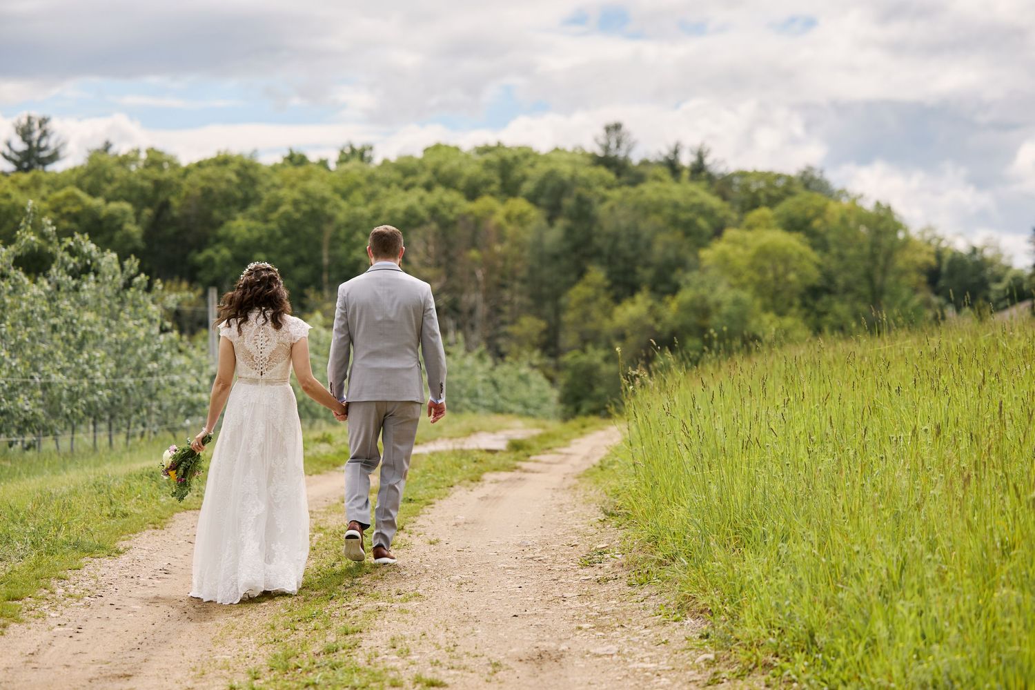 A bride and groom walk hand in hand down a dirt path surrounded by lush green meadows and trees on a summer day.