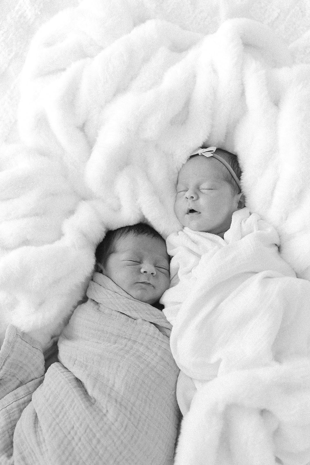 Two sleeping newborns swaddled in white blankets lying side by side on plush white bedding in black and white photography.