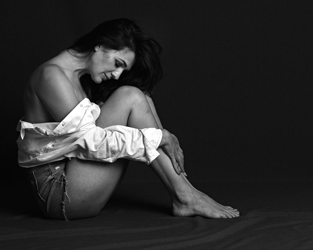 classy boudoir photo of a woman in an open white shirt, sitting on the floor holding her legs.