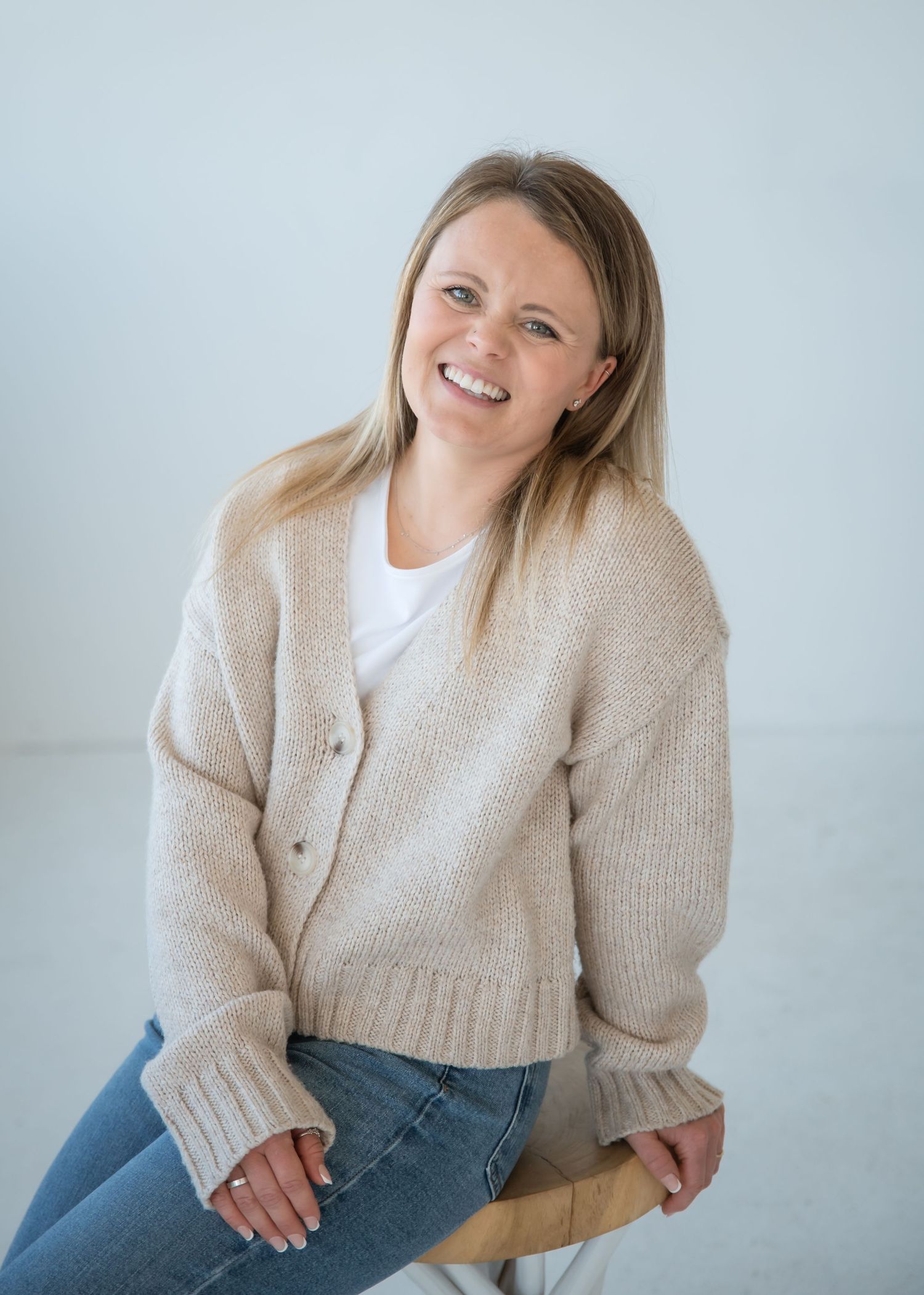 Woman in beige knit sweater and jeans poses casually in studio against light background.
