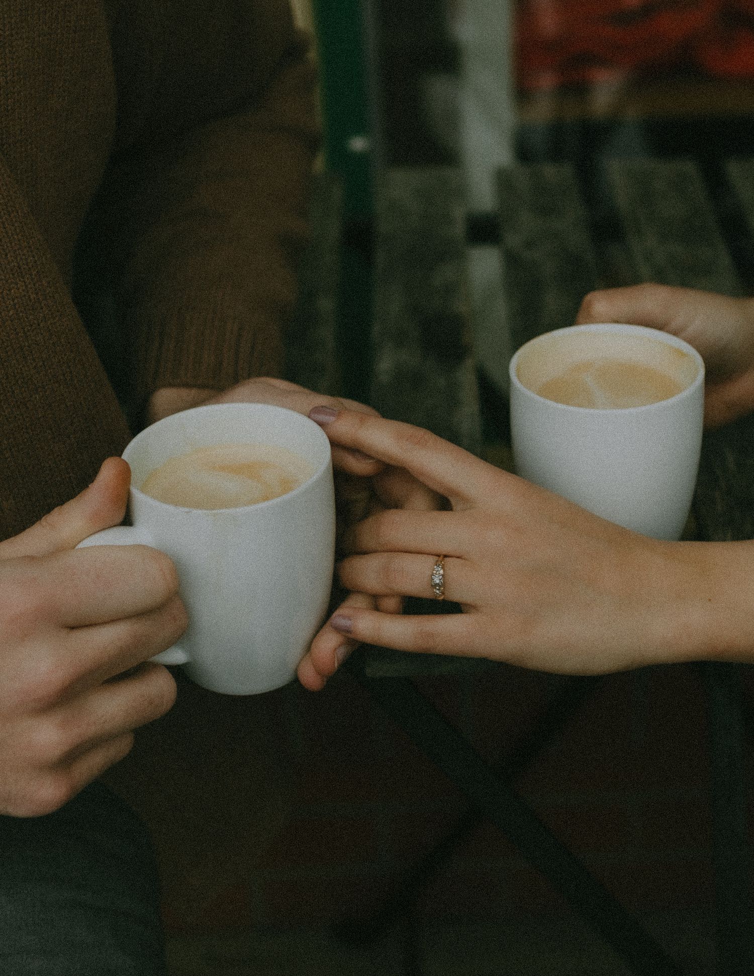 Two people embrace and laugh joyfully while wearing cozy sweaters against an overcast sky and water backdrop.