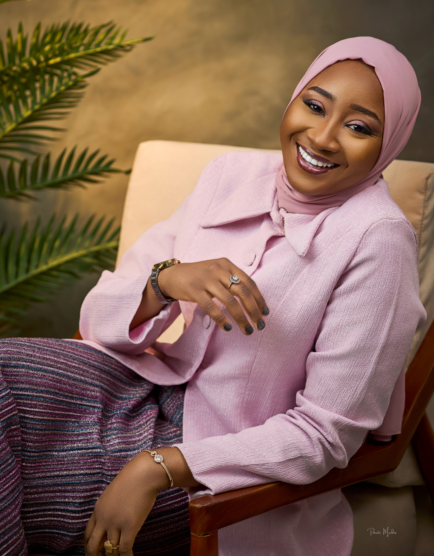 Professional portrait of a woman in pink hijab and blazer sitting in a chair with tropical plants.