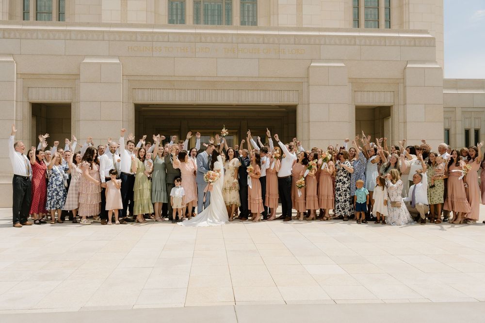 Large group wedding photo with guests in pastel colors gathered outside a light stone building.