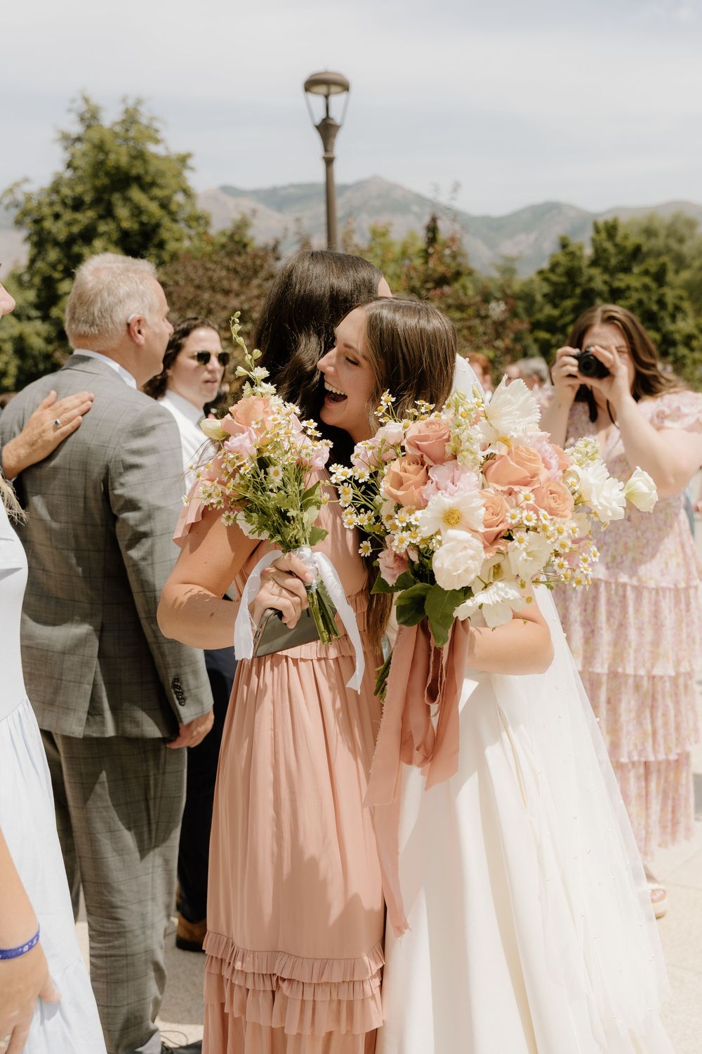 A joyful outdoor gathering with floral bouquets in soft pink and white against a mountain backdrop.