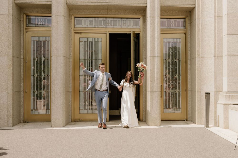 Joyful couple celebrating with raised arms at grand entrance of elegant building with art deco doors.