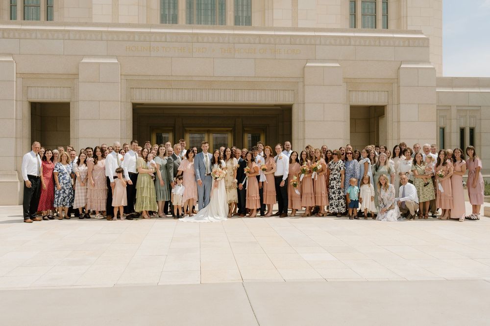 Large wedding group photo with guests in pastel dresses gathered outside a stone building.