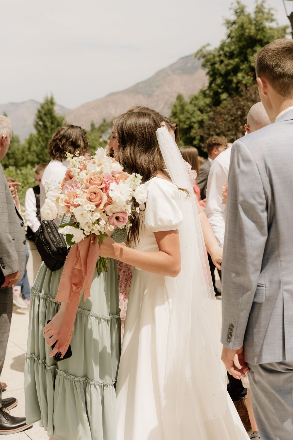 Wedding party gathered outdoors with peach bouquets against a mountain backdrop.