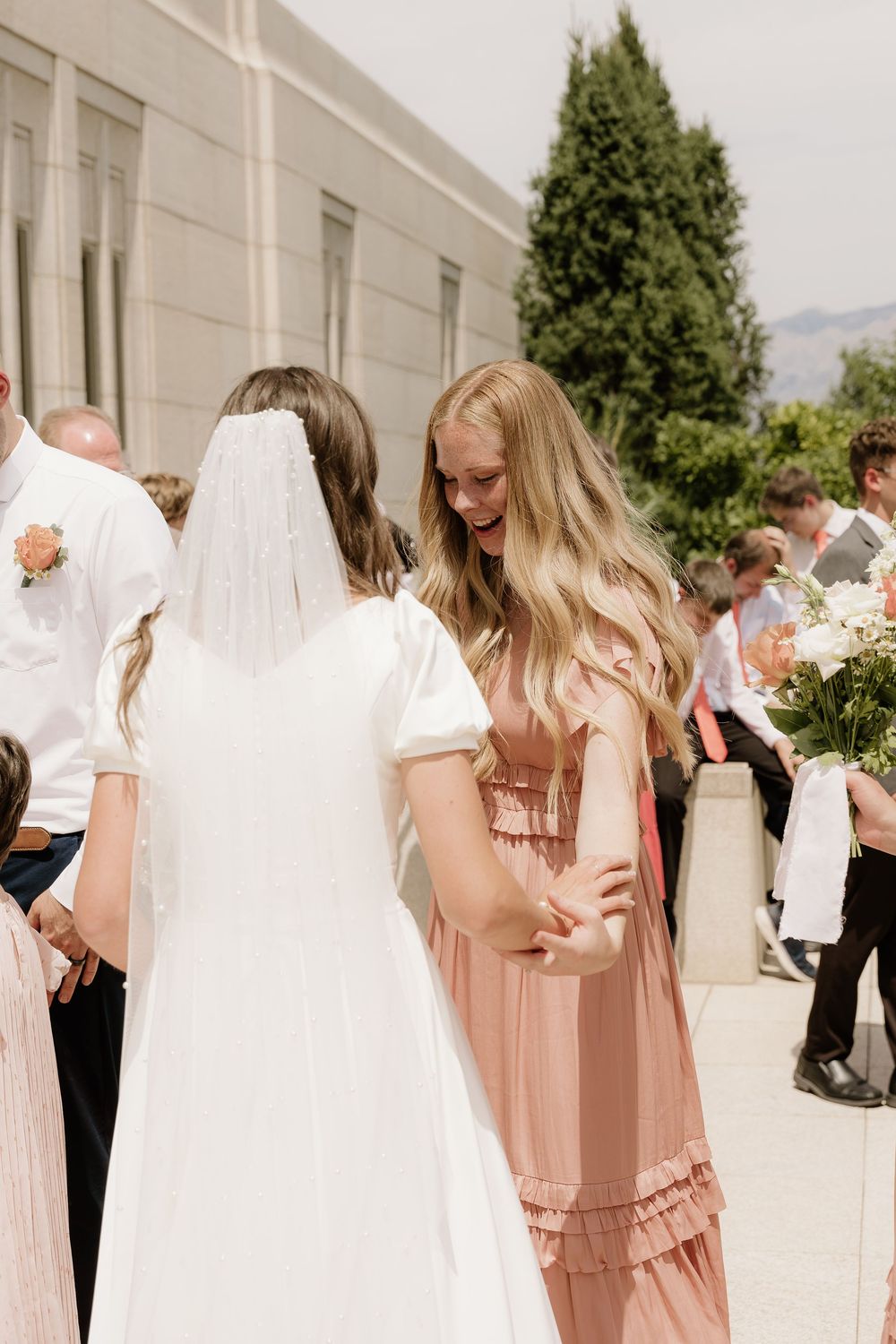 A wedding celebration outside a building with guests, featuring a bride in white and an attendant in blush pink.