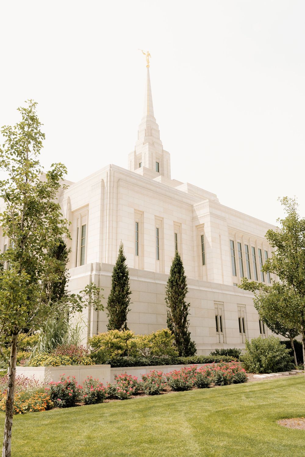 A majestic white Mormon temple with spires and beautiful landscaping featuring cypress trees and flower gardens.