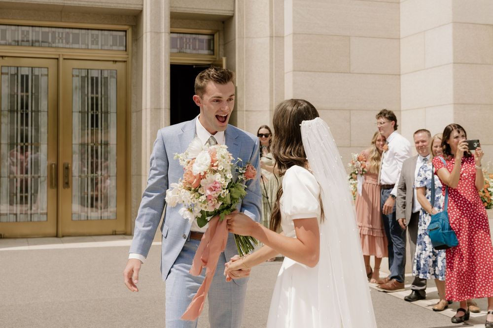 Wedding couple exchanges flowers outside elegant building entrance as guests look on.