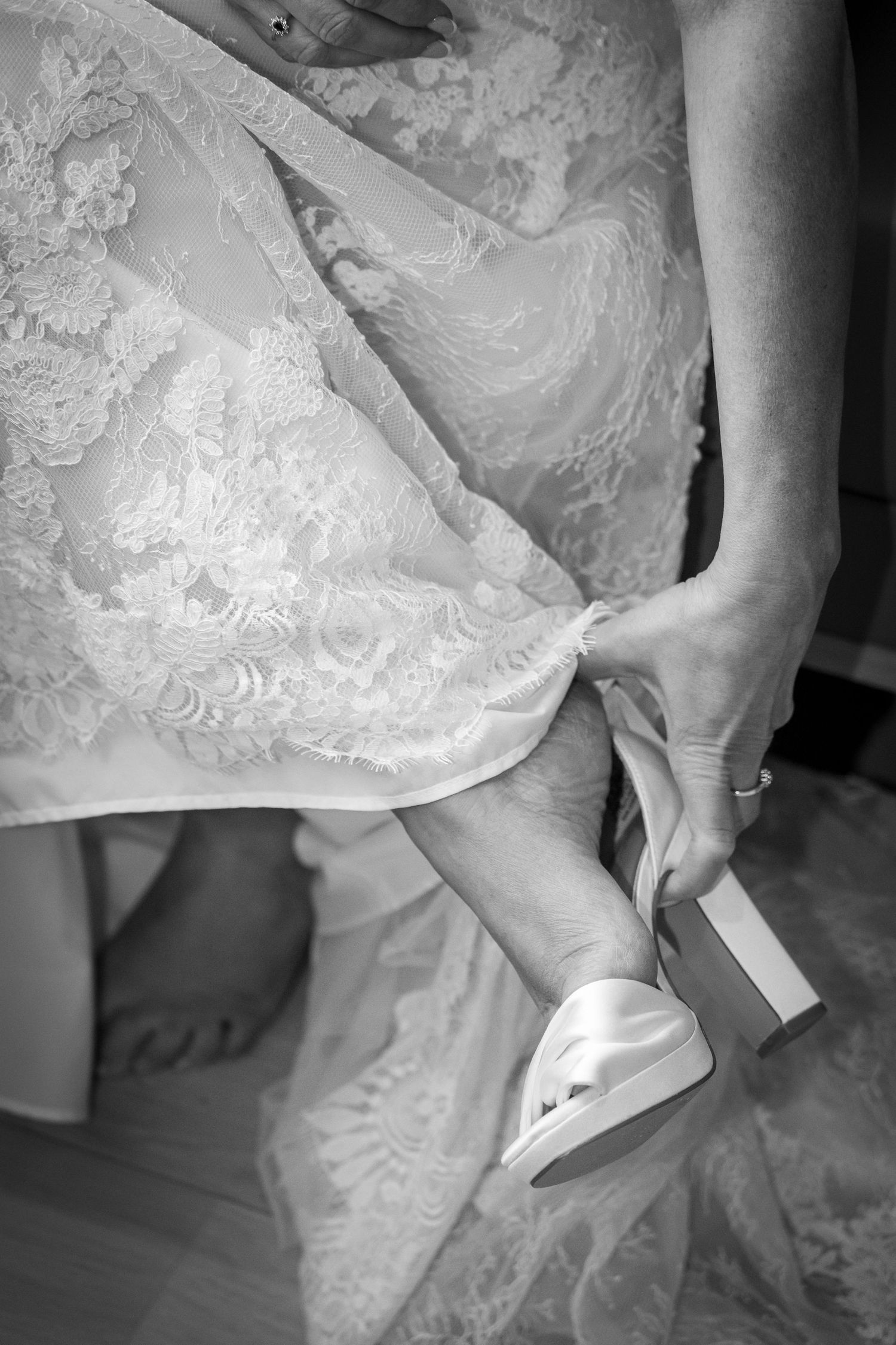 Close-up black and white photo of bride in lace gown having white satin wedding shoe fastened.
