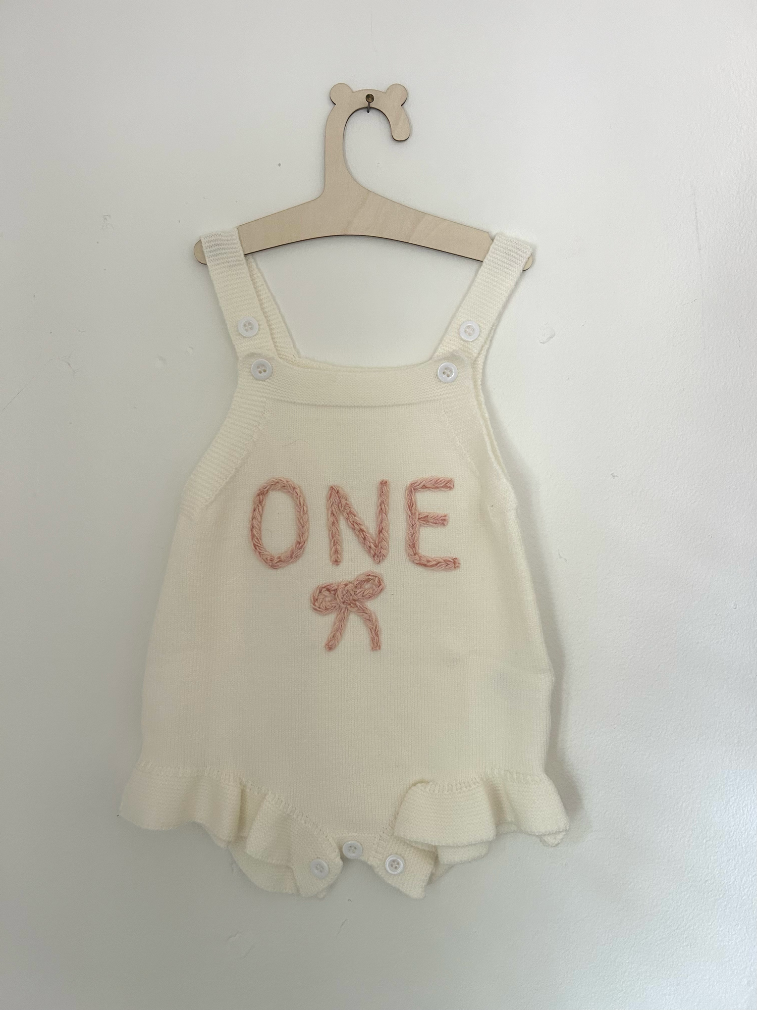 White knit baby romper with 'ONE' text and small bow detail in pink embroidery against hanging clothing background.