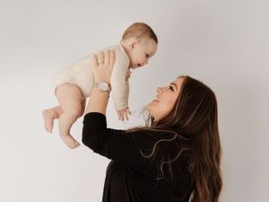 A person in a black sweater lifts a baby wearing a light colored onesie against a white studio backdrop.