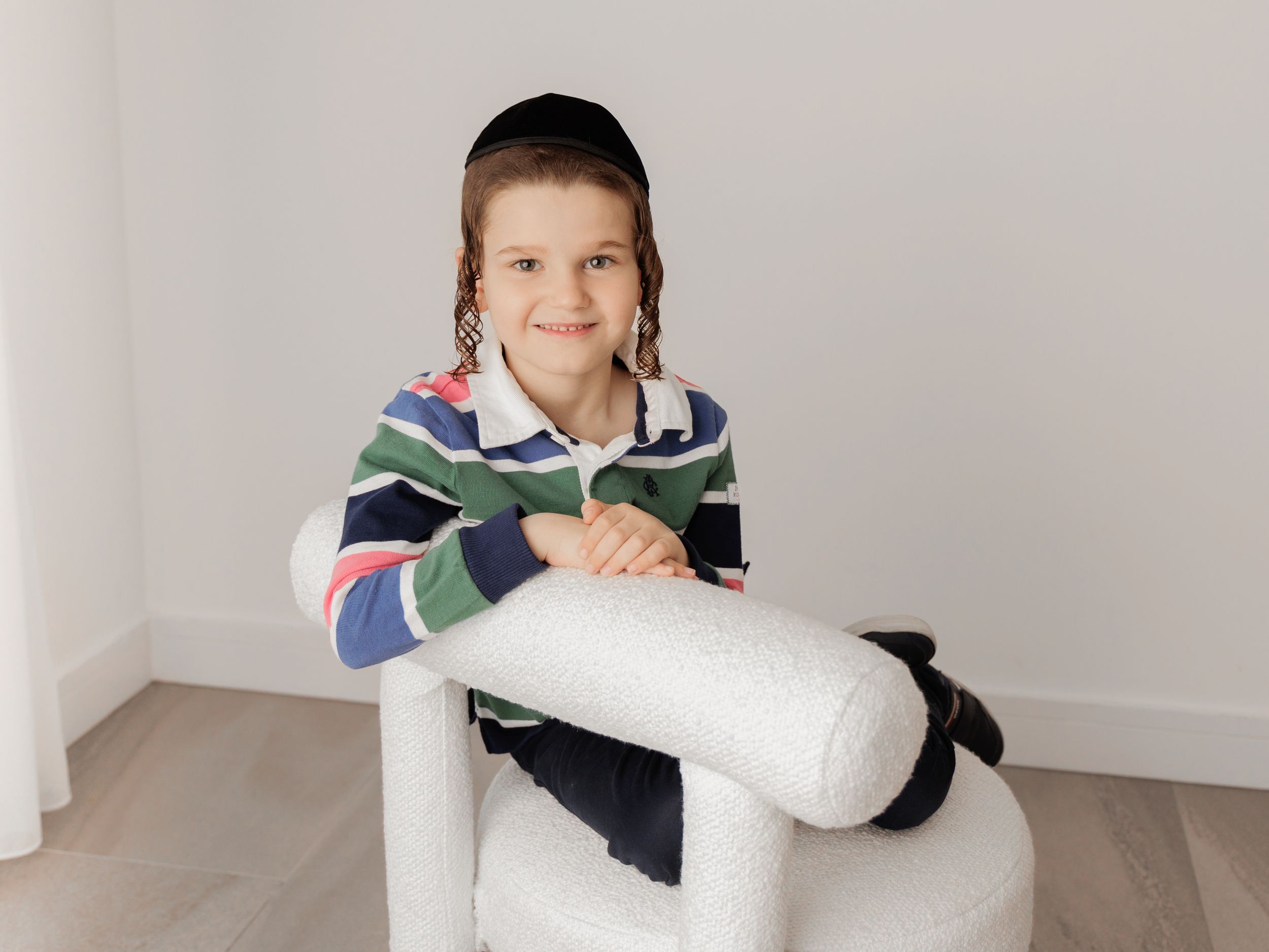 A content young person in a striped hoodie sitting on a white chair in a minimalist room.