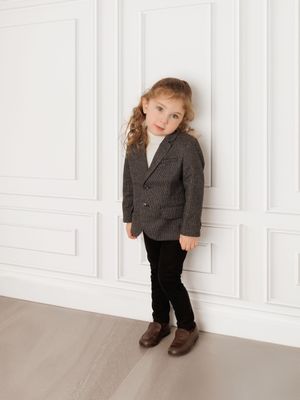 Small child wearing a gray blazer and black pants poses against white paneled wall in classic portrait style.