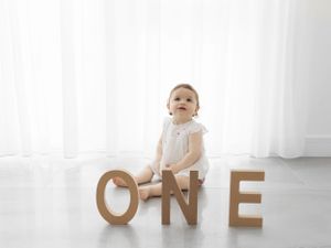 Baby in white outfit sitting next to wooden 'ONE' letters by sheer curtains.