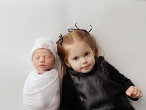 A baby wrapped in white swaddle blanket lying next to a smiling toddler in dark clothing.