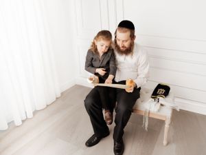 Parent and child sit together reading a book on a white chair in minimalist room setting.