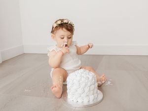 A baby in white sitting next to a white frosted cake during a first birthday cake smash photo session.
