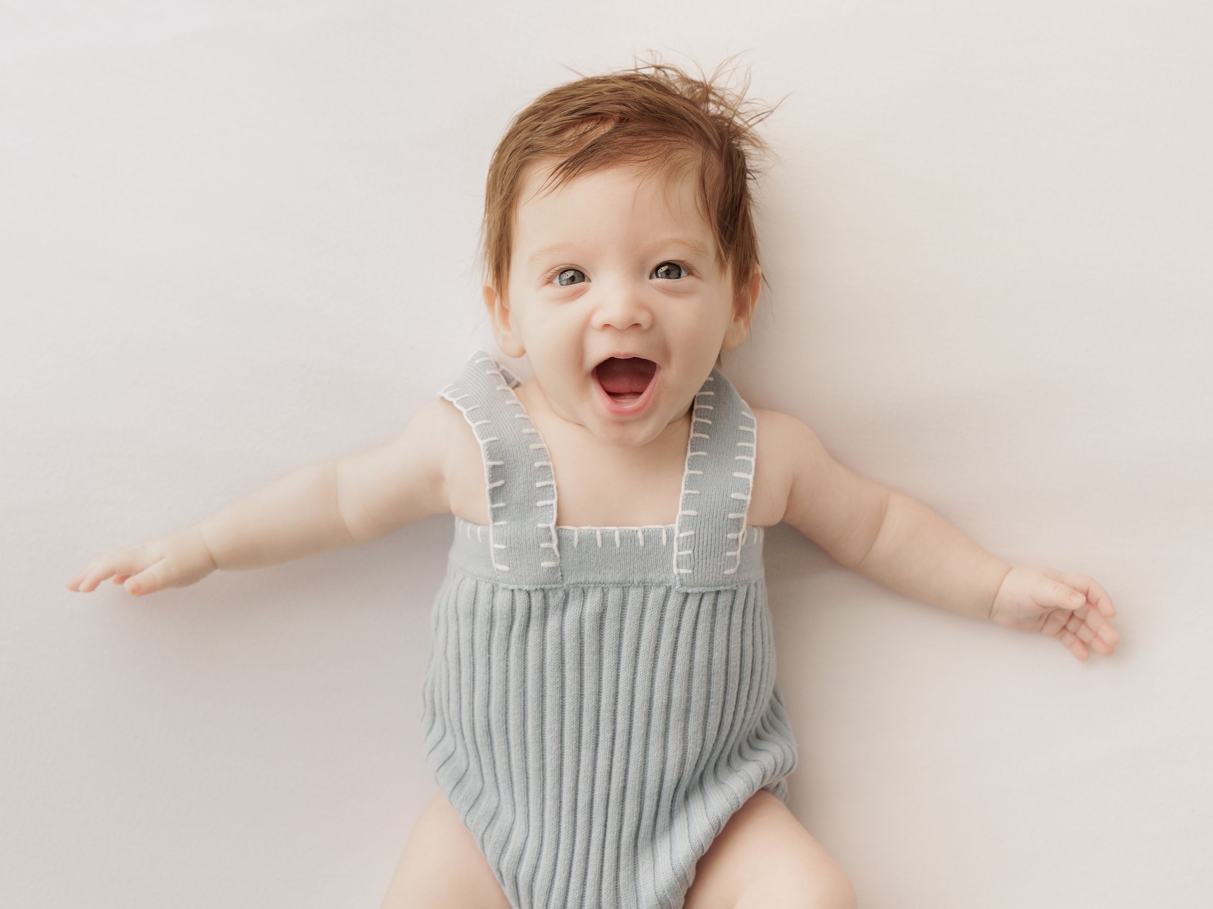 Baby wearing light blue striped romper lies on white blanket with joyful expression and outstretched arms.
