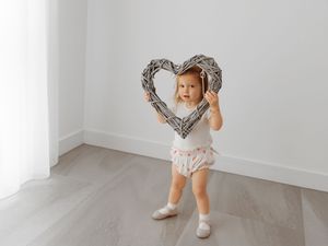 A child in light colored clothing holds up a decorative heart-shaped frame against a white wall.