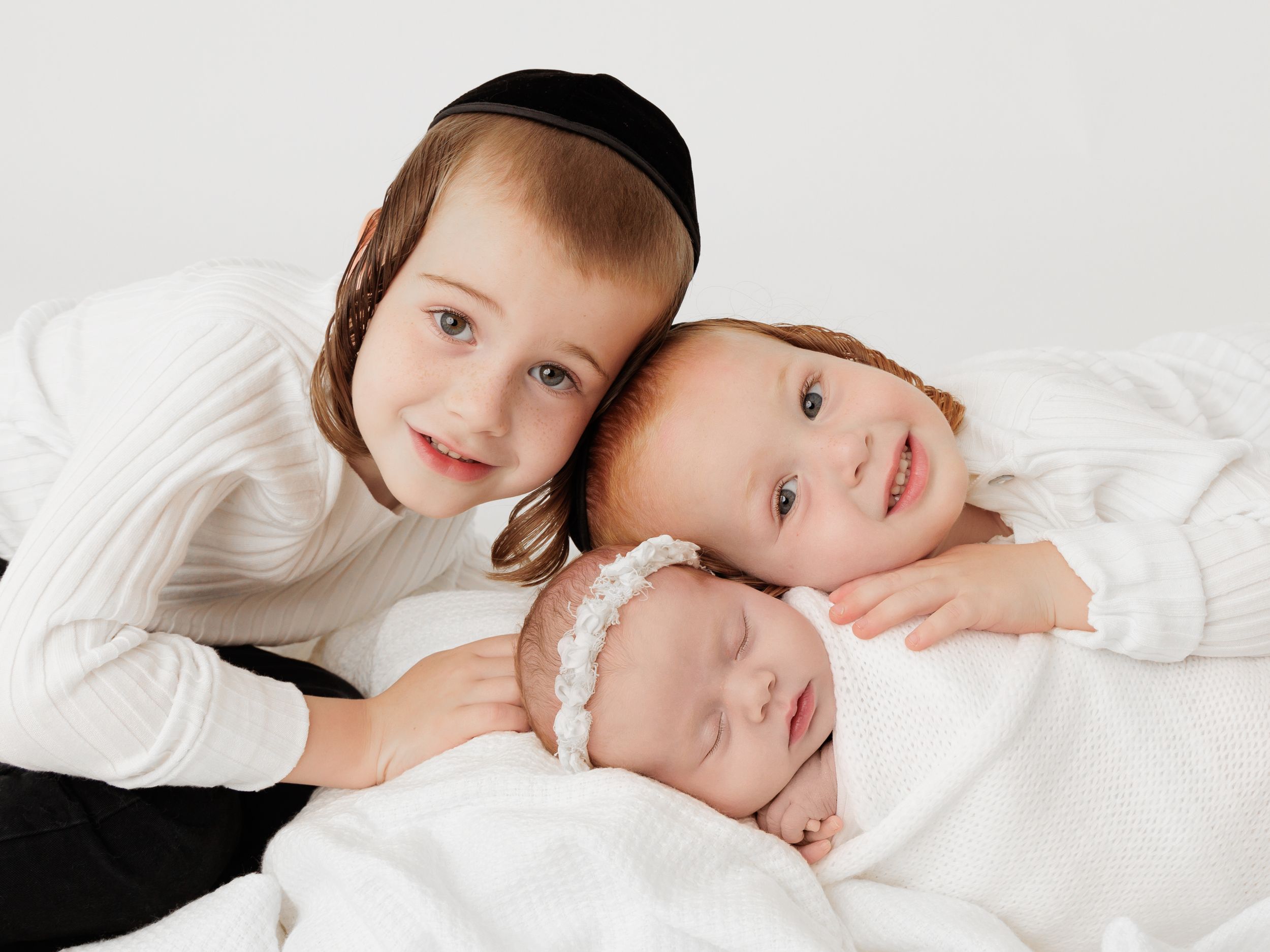 Three children in white clothing lying together on white bedding in a sweet candid moment.