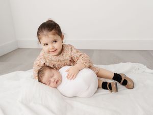 Two siblings on white floor with older child gently embracing sleeping newborn baby in soft lighting.