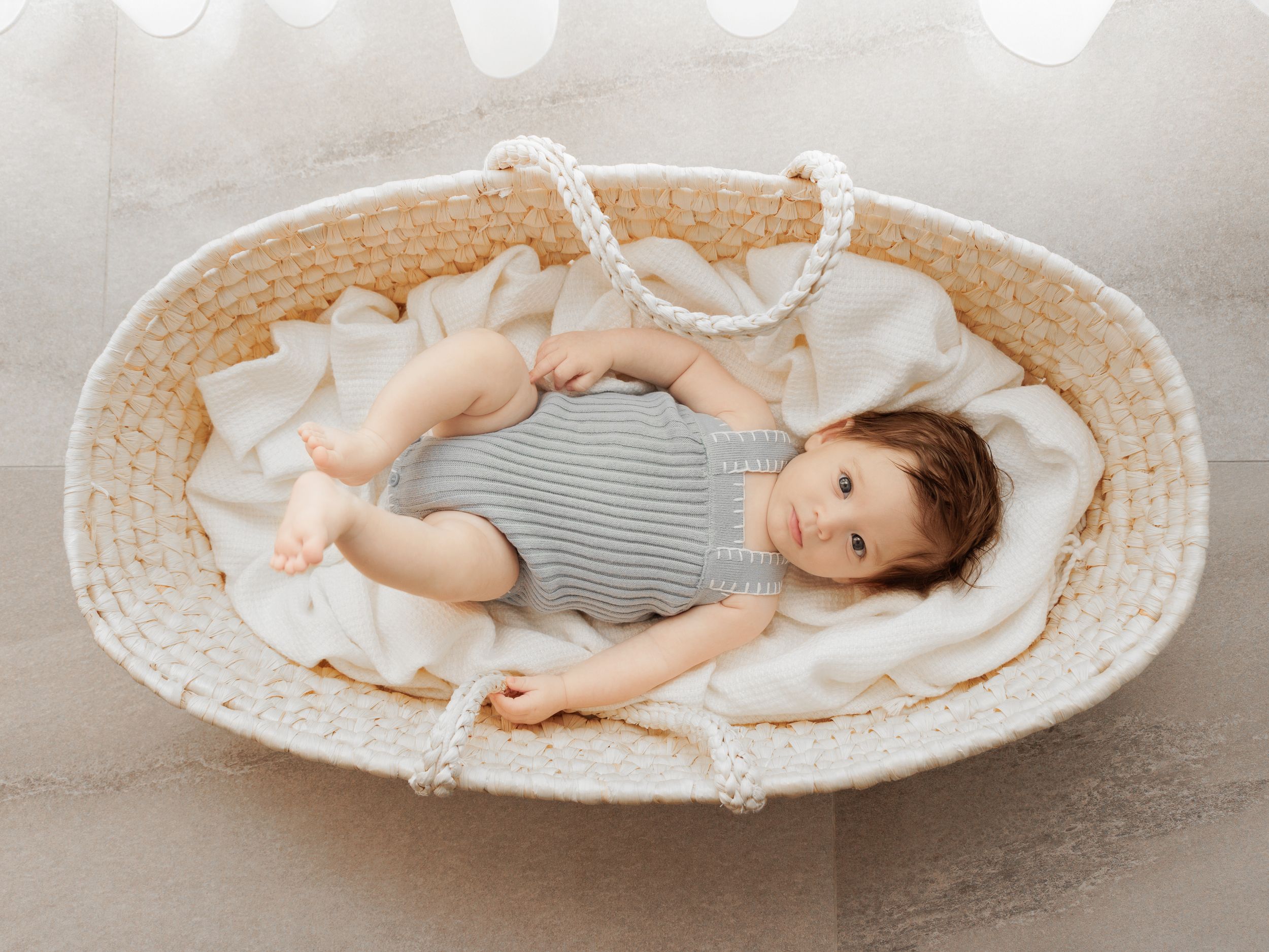 Baby lying in a round woven basket wearing a gray striped outfit and white blanket.