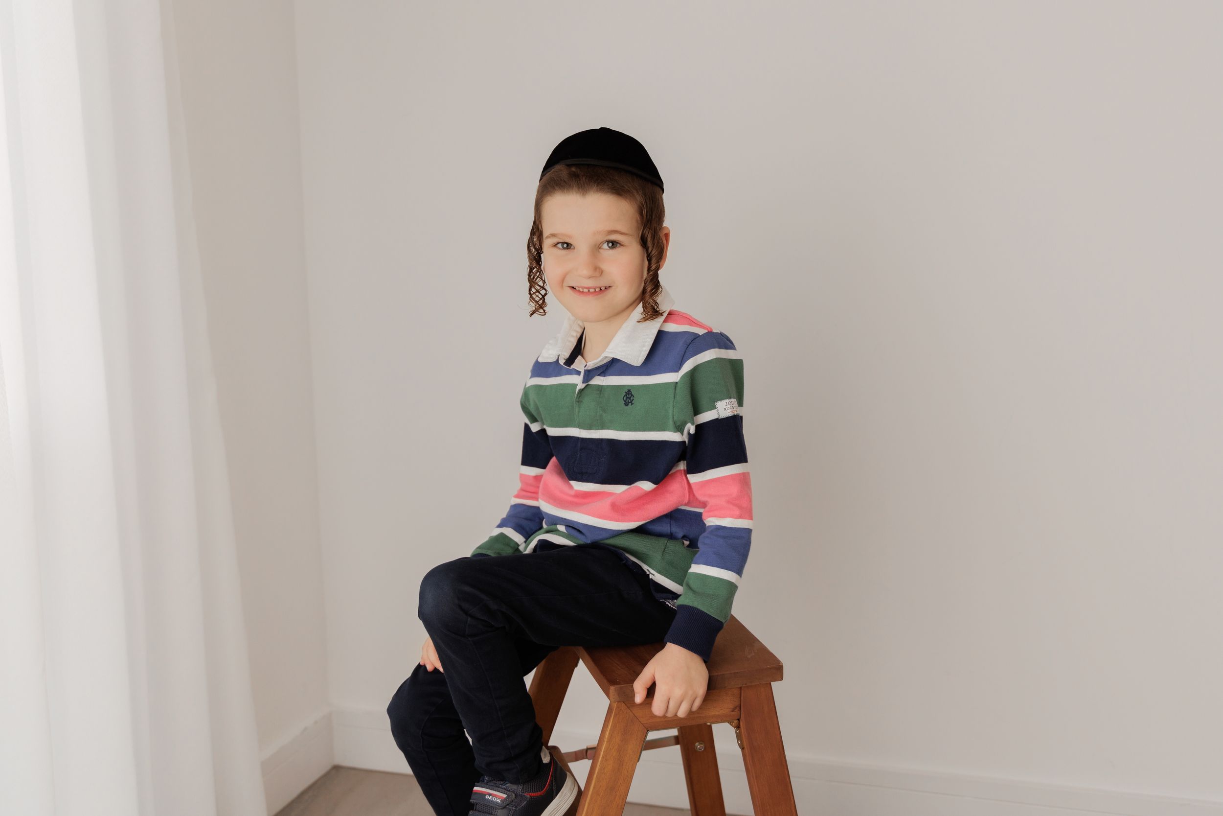A child in a striped multicolor sweater sits on a wooden stool against a white wall.