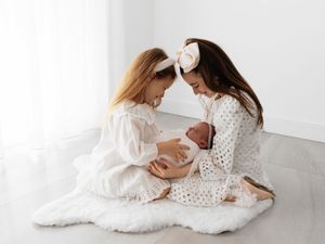Two people in white dresses sitting on a fluffy rug share a tender moment with a newborn baby.