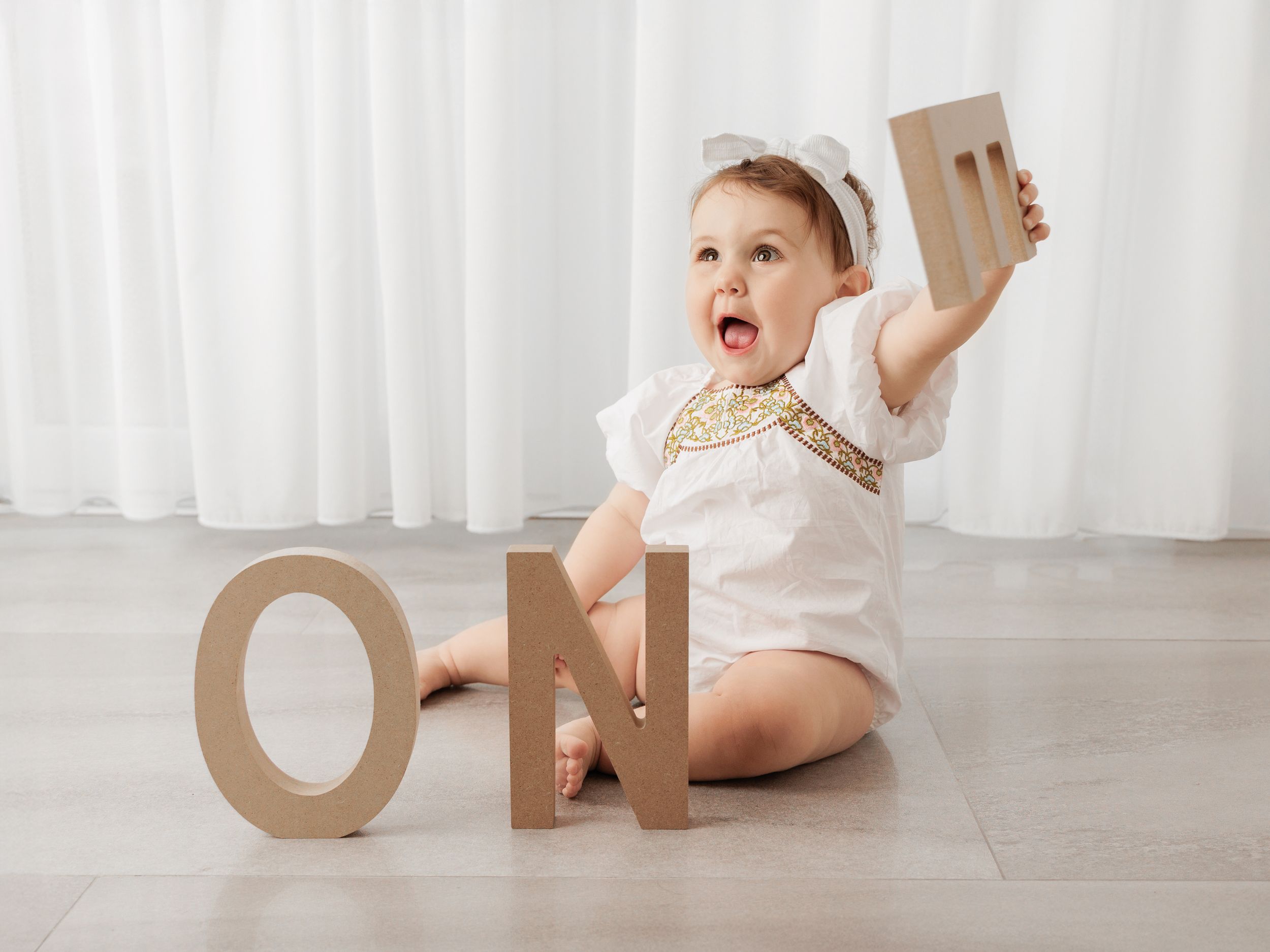 Excited baby in white outfit sits on floor with decorative 'ONE' wooden numbers for first birthday celebration.