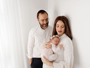 A couple dressed in white smiles lovingly while holding their newborn baby against a white background.