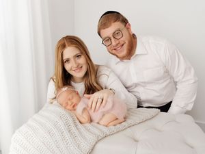 A couple in white attire cuddles with their newborn baby on white bedding.