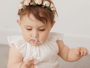 A small child wearing a white dress and floral crown tastes frosting during what appears to be a cake smash photo session.