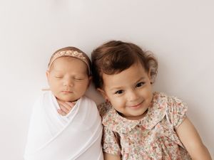 A baby wrapped in white swaddle and an older sibling in a floral dress pose together for a portrait.
