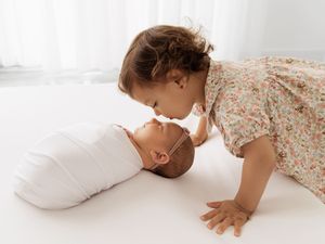 Two young children in floral dress interact sweetly together on white bedding in bright natural light.