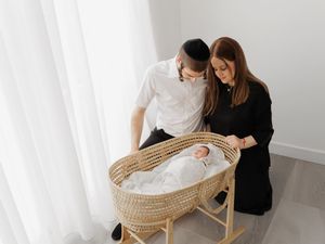 Couple lovingly gazing at sleeping baby in woven bassinet with white curtains in background.